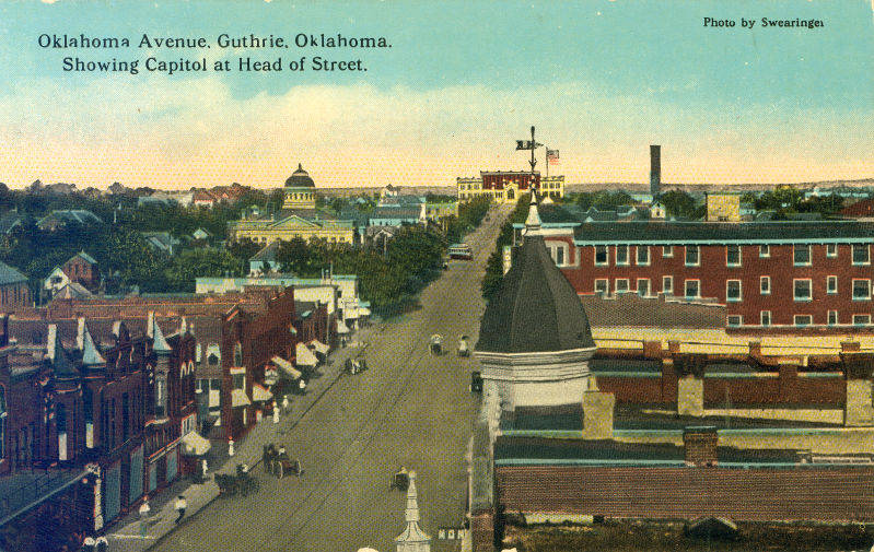 Several blocks of brick buildings along Oklahoma Avenue in Guthrie, Oklahoma are shown in this color postcard. Oklahoma's original Capitol building is at the head of the street.