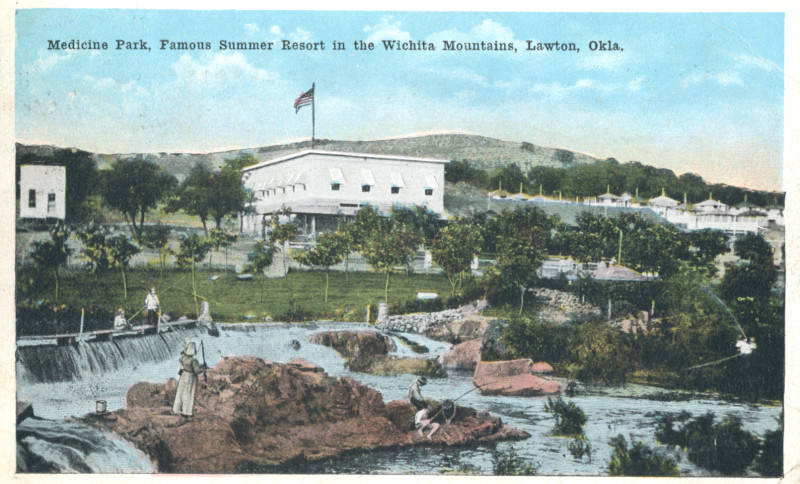 A vintage postcard-style image of Medicine Park, Oklahoma, featuring a scenic view of the area. The image shows a flowing stream with small waterfalls and people fishing. In the background, a large, white building with a flat roof and a flagpole flying the American flag sits atop a hill. The sky is a light blue with a few white clouds.